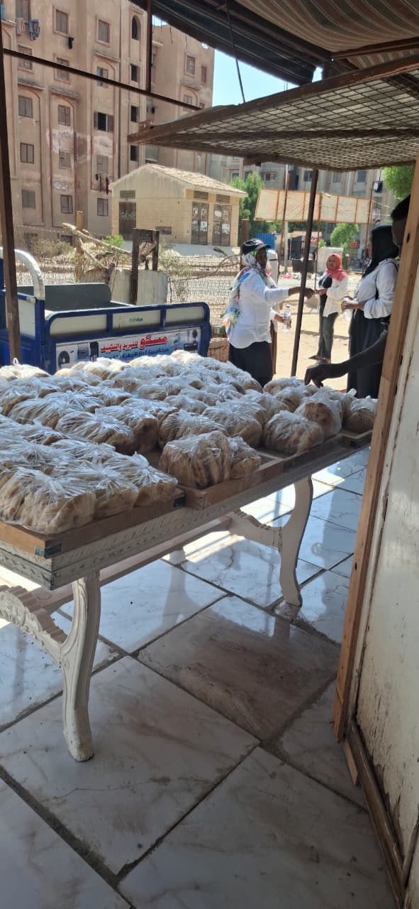 Trays of bagged bread on a table beside a loaded delivery cart, with volunteers alongside.